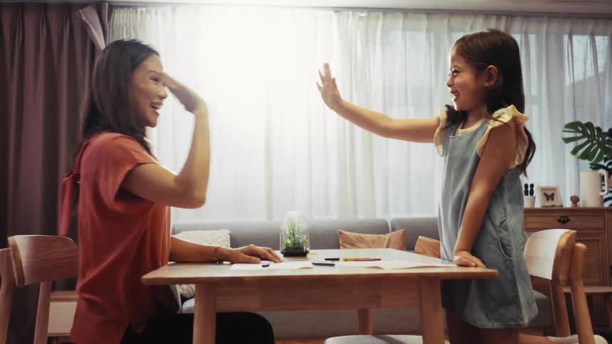Happy Asian mother and her adorable daughter have a creative drawing session, ending with a joyful high-five in their cozy living room