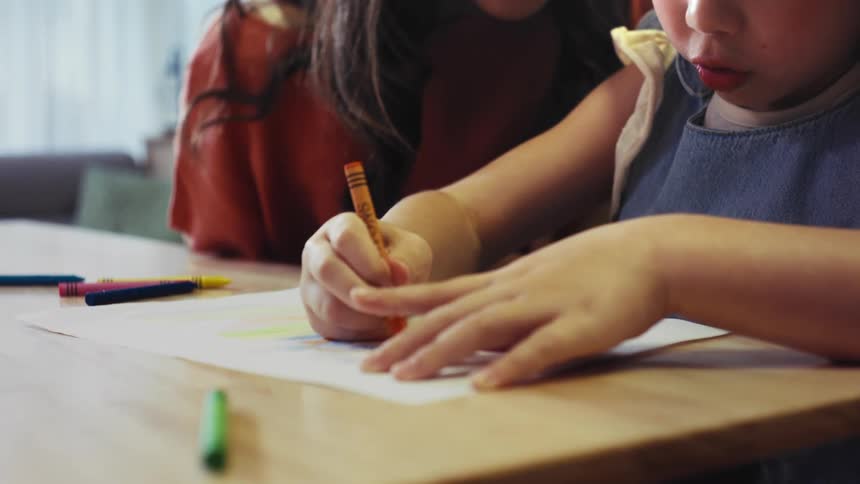 A loving mother watches her young daughter as she concentrates on coloring a picture with crayons at a wooden table