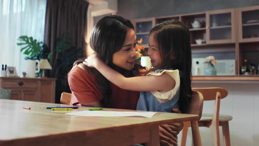 A young daughter gives her loving mother a big, warm hug at the dining table in their home in a heartwarming slow motion clip