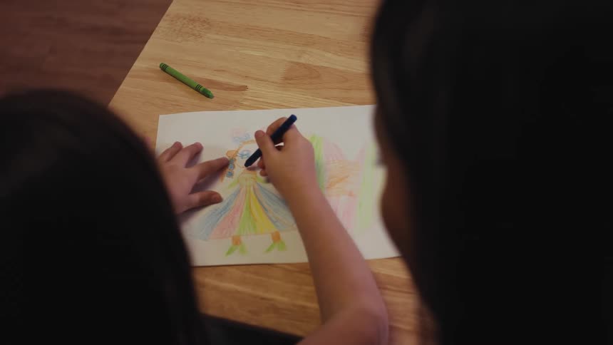 An over-the-shoulder shot captures a young girl coloring a vibrant picture of a princess on a wooden table