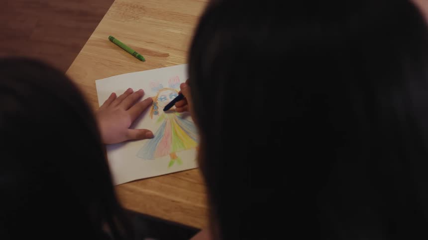 An over-the-shoulder shot captures a young girl coloring a vibrant picture of a princess on a wooden table