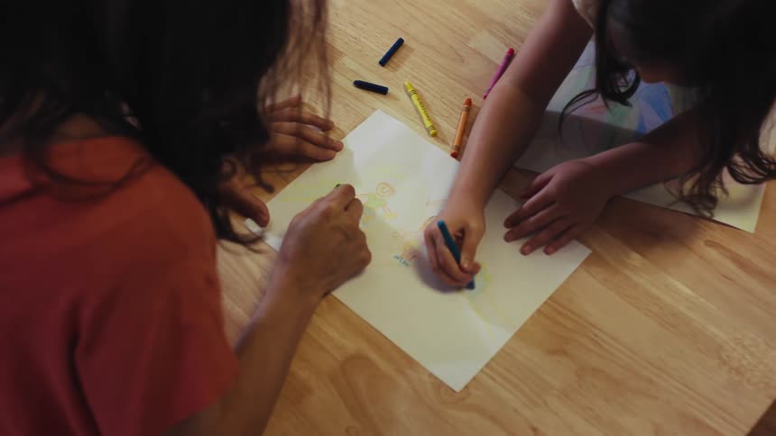 An overhead shot of a mother and daughter coloring together with crayons at a wooden table