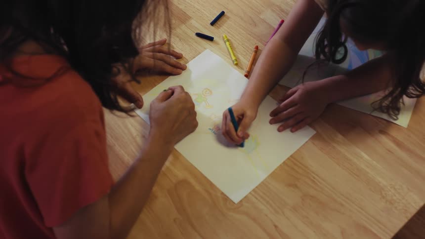 An overhead shot of a mother and daughter coloring together with crayons at a wooden table