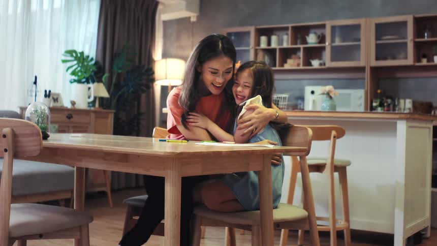 A loving mother and her adorable young daughter share a heartwarming hug while drawing together at the dining table in their cozy home