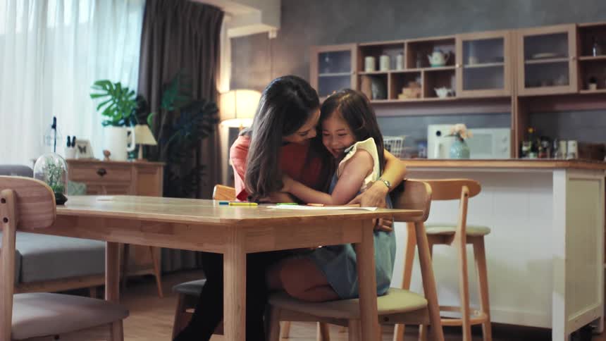 A loving mother and her adorable young daughter share a heartwarming hug while drawing together at the dining table in their cozy home