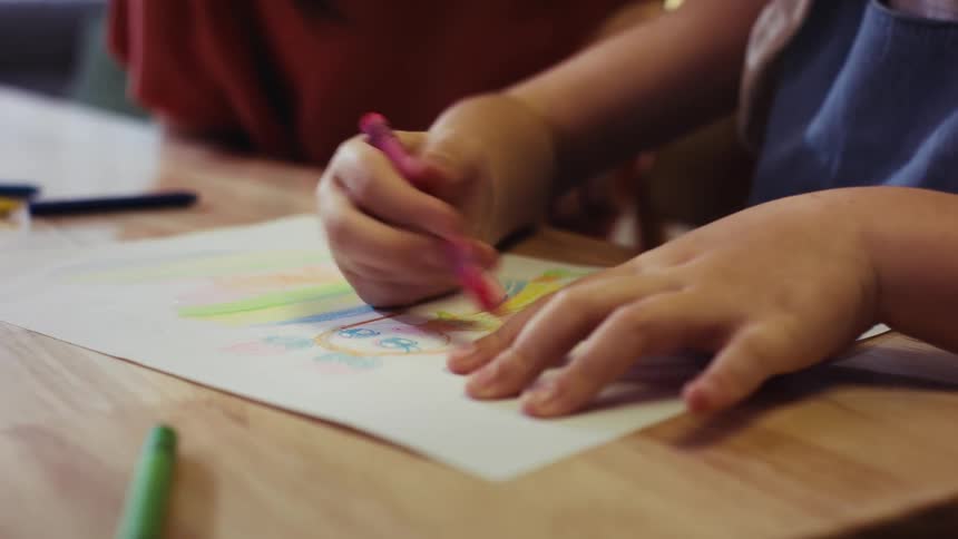 A close-up of a young child's hands carefully coloring a drawing with a pink crayon on a wooden table