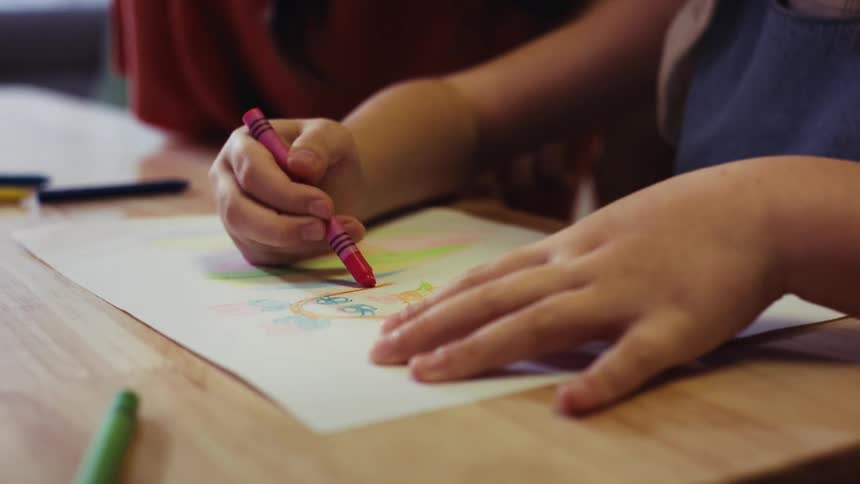 A close-up of a young child's hands carefully coloring a drawing with a pink crayon on a wooden table