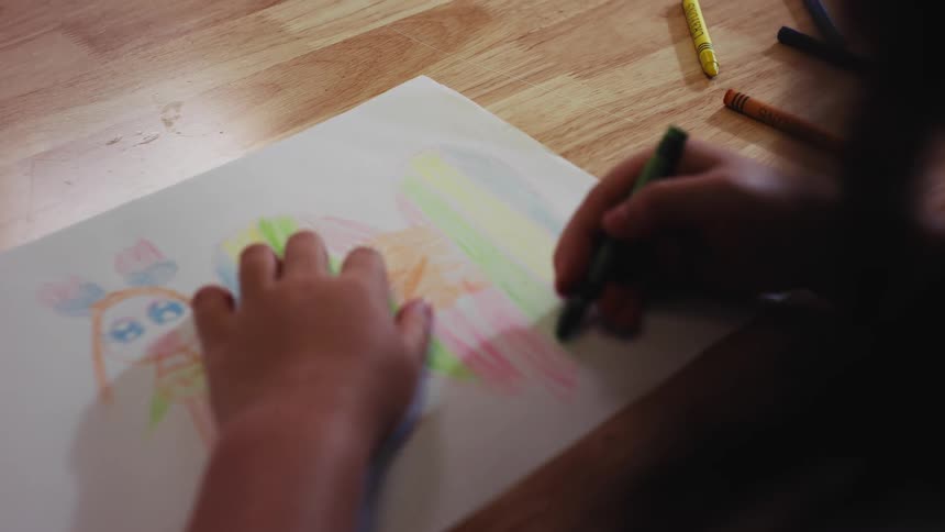 Over-the-shoulder close-up of a young child coloring a picture of a girl and a rainbow heart with crayons