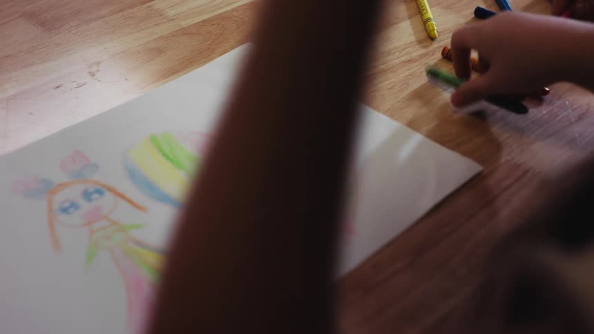 Over-the-shoulder close-up of a young child coloring a picture of a girl and a rainbow heart with crayons