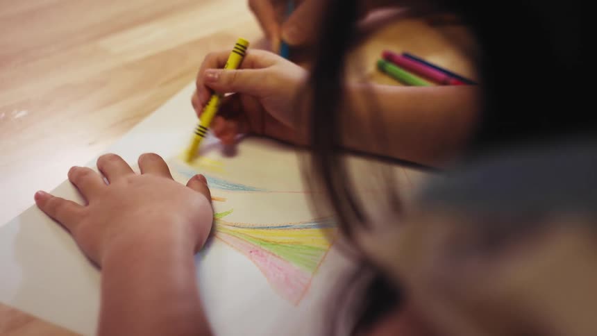 An over-the-shoulder close-up shot of two people coloring a rainbow together with crayons on a wooden surface