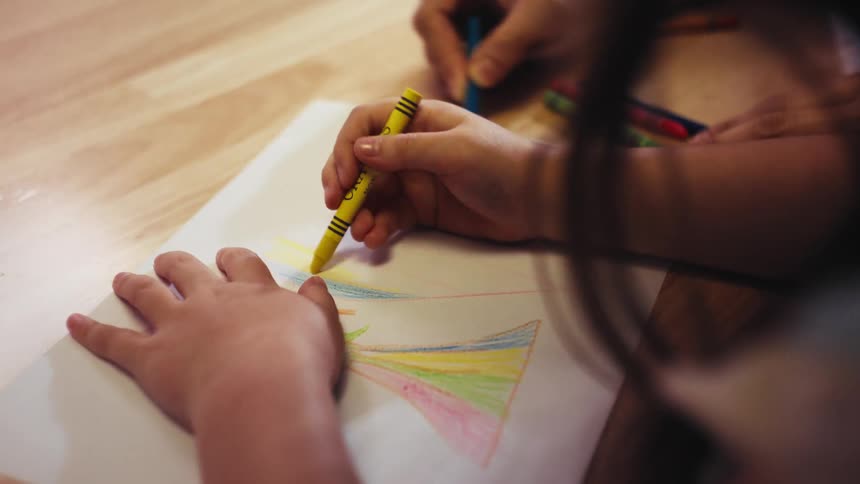 An over-the-shoulder close-up shot of two people coloring a rainbow together with crayons on a wooden surface