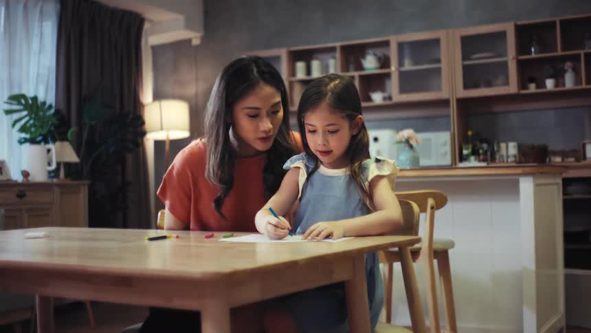 An Asian mother and her cute daughter spend a happy evening together, drawing with crayons at the dining table and sharing a loving hug