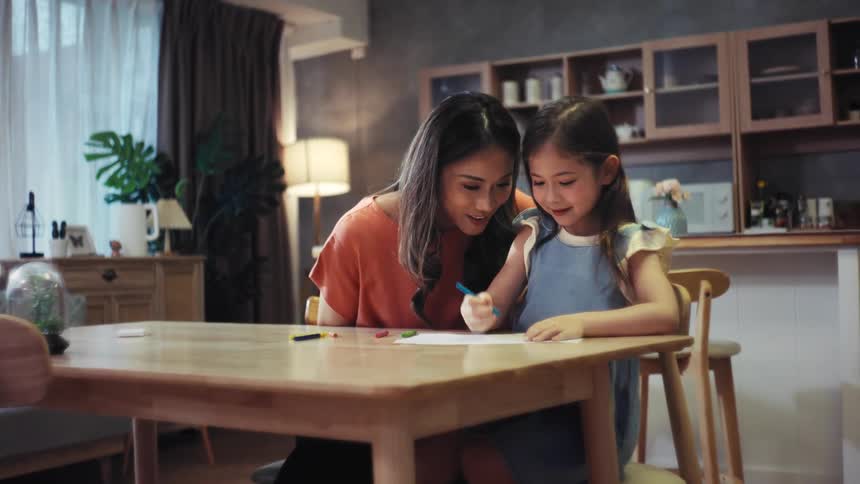 An Asian mother and her cute daughter spend a happy evening together, drawing with crayons at the dining table and sharing a loving hug