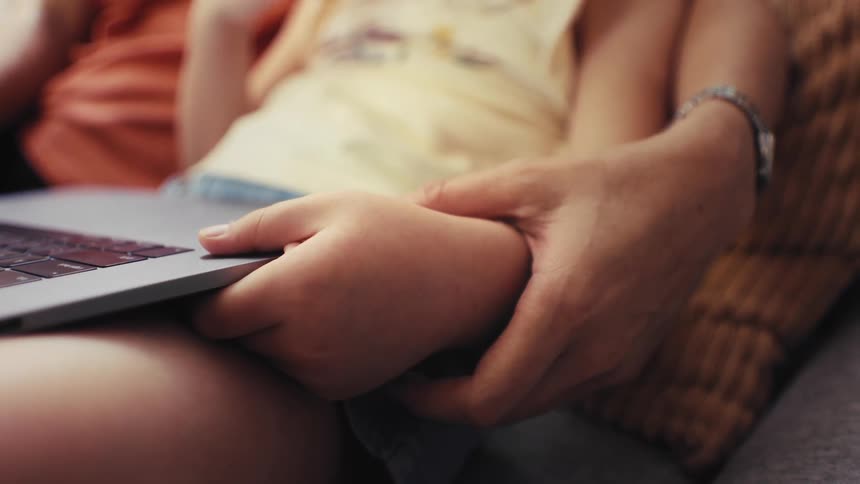 A tender close-up shows a parent's hand lovingly holding their child's hand while they look at a laptop together