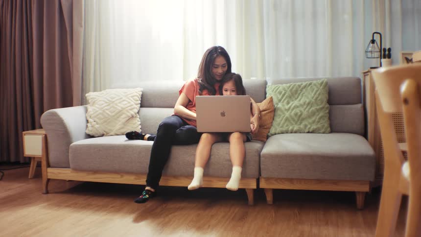 A smiling mother and her young daughter sit together on the couch in their living room, looking at a laptop