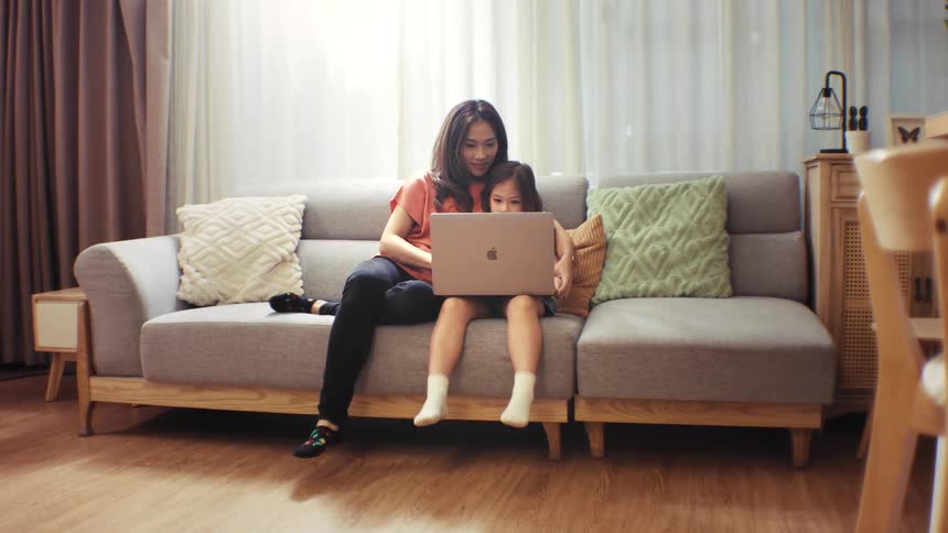 A smiling mother and her young daughter sit together on the couch in their living room, looking at a laptop