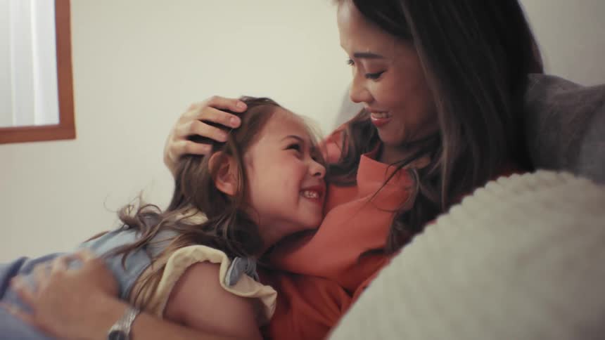 A loving mother and her happy young daughter share an affectionate cuddle on the sofa at home, smiling at each other