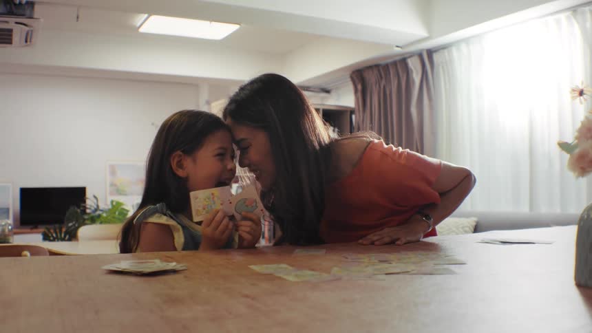An adorable young girl and her loving mother share a joyful moment playing a card game at a wooden table in their bright home