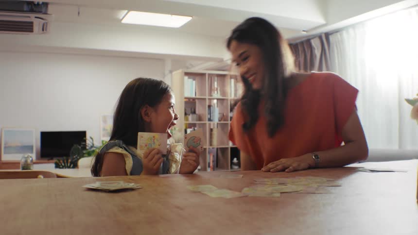 An adorable young girl and her loving mother share a joyful moment playing a card game at a wooden table in their bright home