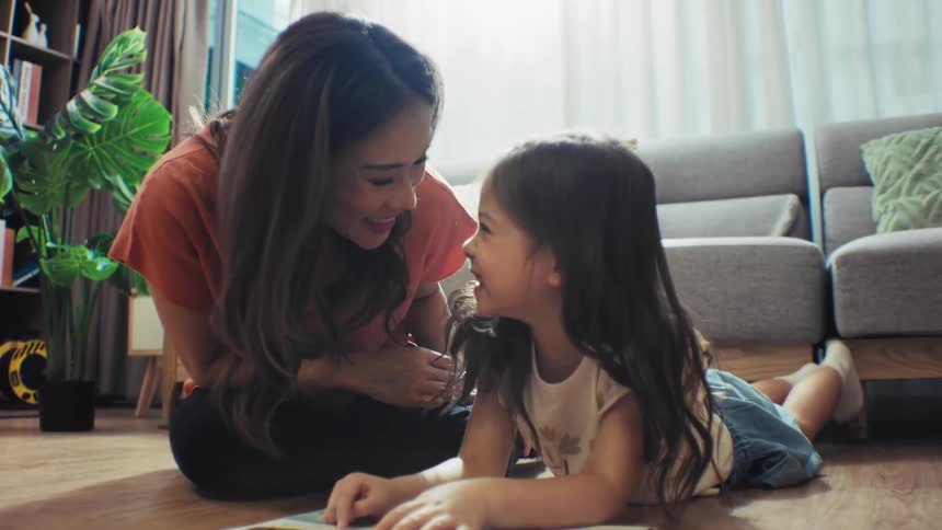 A happy Asian mother reads a storybook with her young daughter, sharing a playful and tender moment on the floor at home