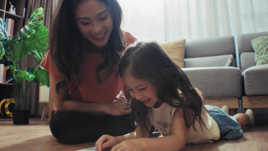 A happy Asian mother reads a storybook with her young daughter, sharing a playful and tender moment on the floor at home