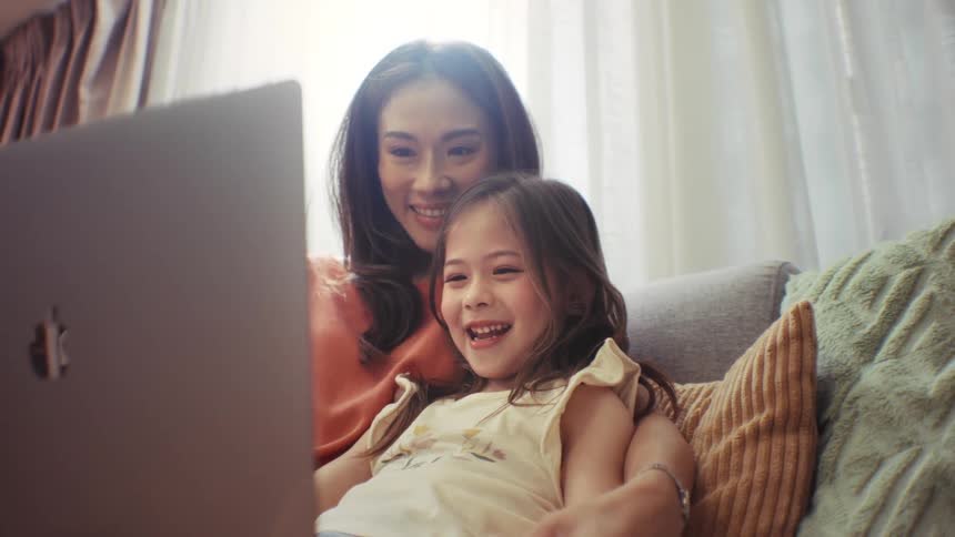 A happy mother and her young daughter laugh while watching an engaging video on a laptop together on the couch at home