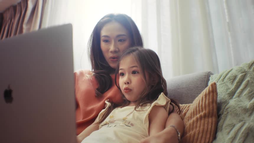 A happy mother and her young daughter laugh while watching an engaging video on a laptop together on the couch at home