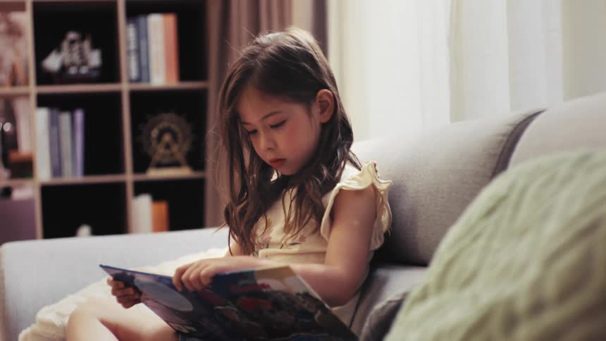 A young girl with long hair sits on a comfy couch, completely absorbed in reading a colorful picture book in her living room