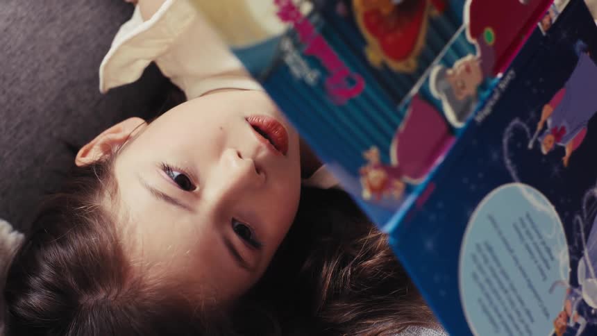 A vertical overhead shot shows a little girl with brown hair lying down while reading a children's storybook