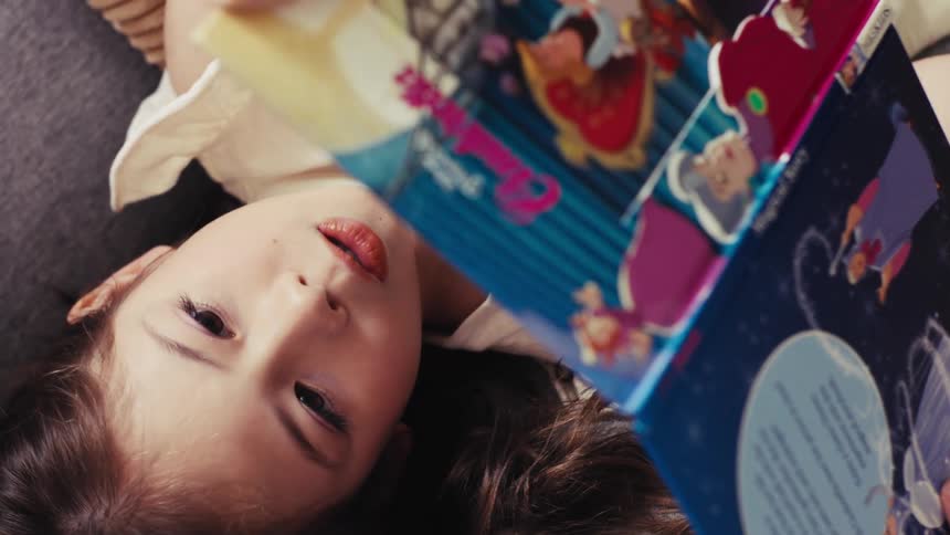 A vertical overhead shot shows a little girl with brown hair lying down while reading a children's storybook