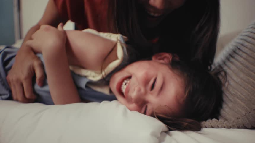 A happy young girl laughs uncontrollably as her mother tickles her while playing together on a bed