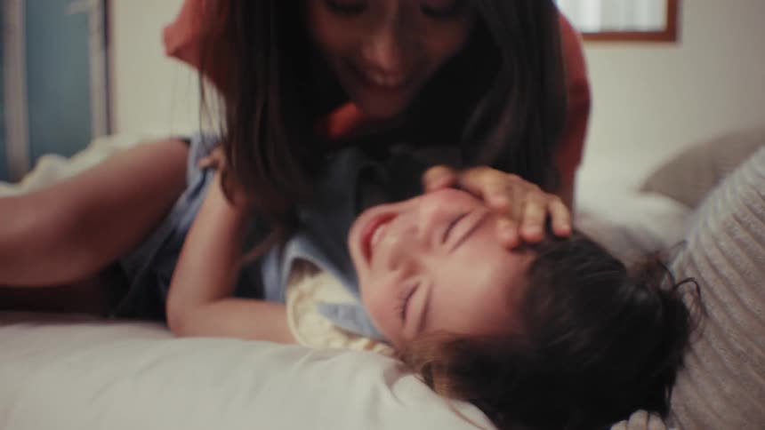 A happy young girl laughs uncontrollably as her mother tickles her while playing together on a bed
