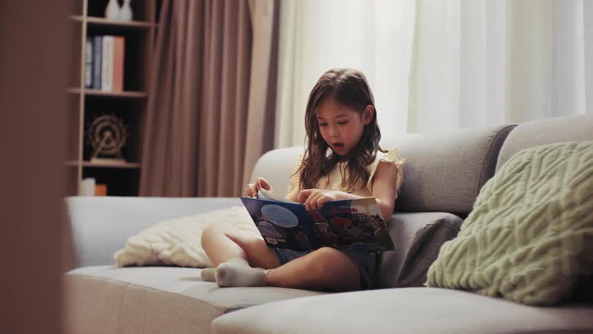 A curious young girl with long hair sits comfortably on a sofa in a bright living room, engrossed in reading a picture book