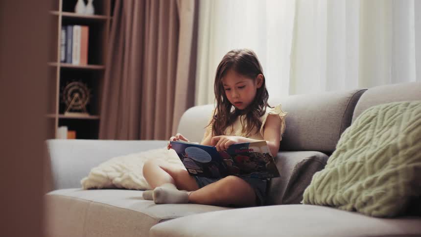 A curious young girl with long hair sits comfortably on a sofa in a bright living room, engrossed in reading a picture book