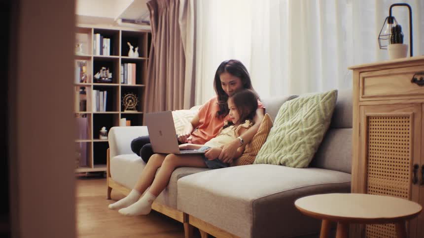 A loving mother and her daughter cuddle on the couch while using a laptop together in their bright and cozy living room