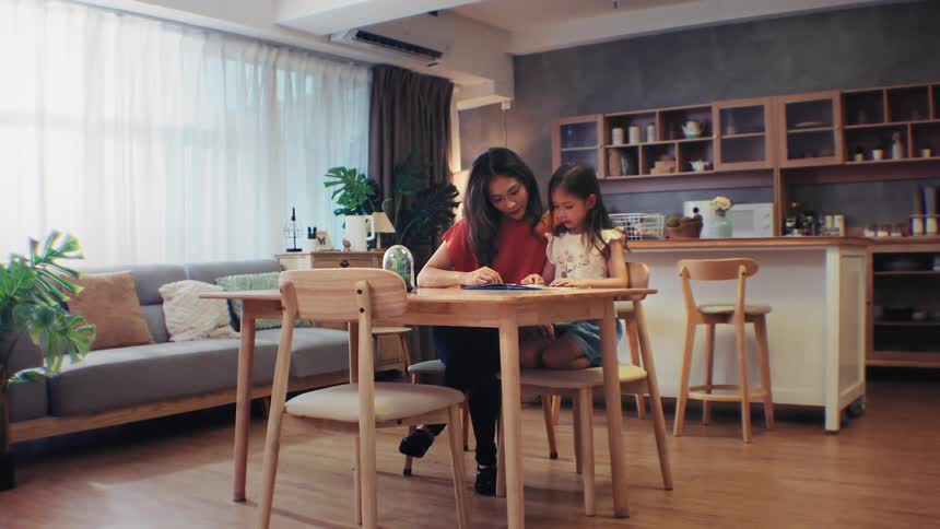 A loving mother sits with her young daughter at the dining table, helping her with schoolwork in their modern home