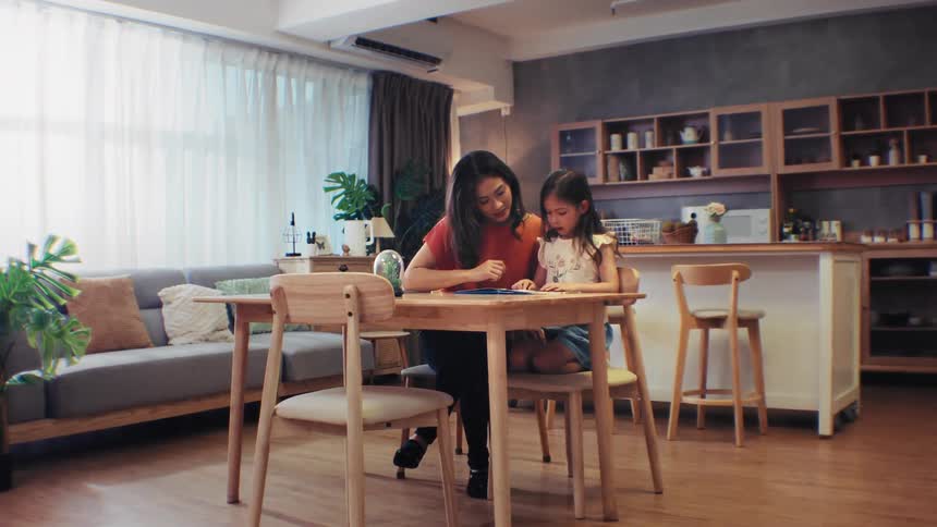 A loving mother sits with her young daughter at the dining table, helping her with schoolwork in their modern home