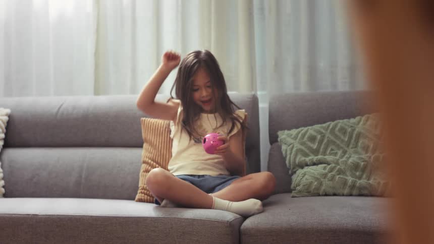 An adorable little girl sitting on the sofa joyfully celebrates a win while playing with a pink toy