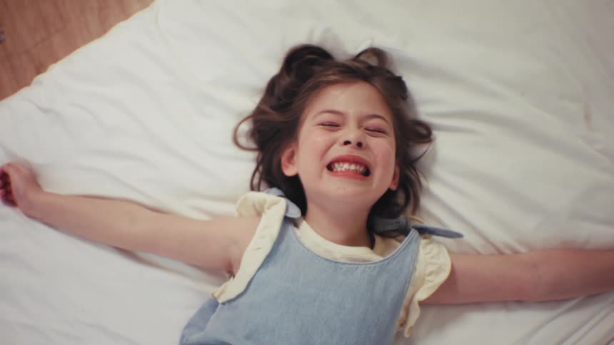 Top-down cinemagraph of a cute little girl smiling and then pretending to sleep on a white bed