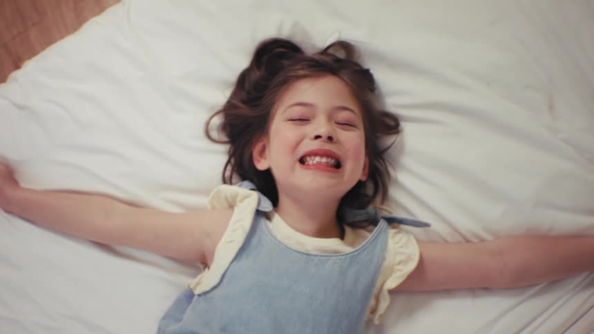 Top-down cinemagraph of a cute little girl smiling and then pretending to sleep on a white bed