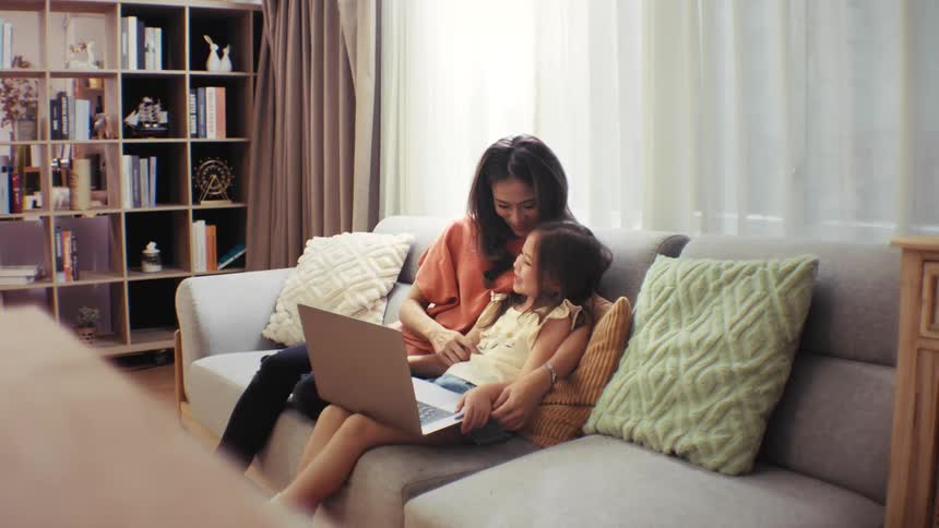 A loving Asian mother and her young daughter bond while using a laptop together on the sofa in their cozy living room