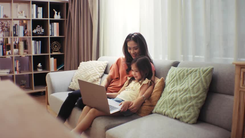 A loving Asian mother and her young daughter bond while using a laptop together on the sofa in their cozy living room
