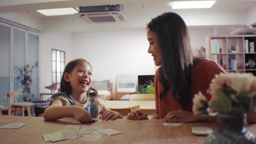 A joyful young girl laughs and smiles while playing a fun card game with her loving mother at the dining table in their home