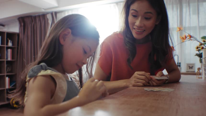 A young Asian mother and her cute daughter play a card game together at a table in their sunlit home