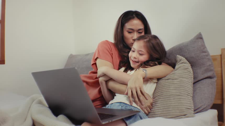 An affectionate mother and her happy young daughter cuddle together while watching a movie on a laptop in bed