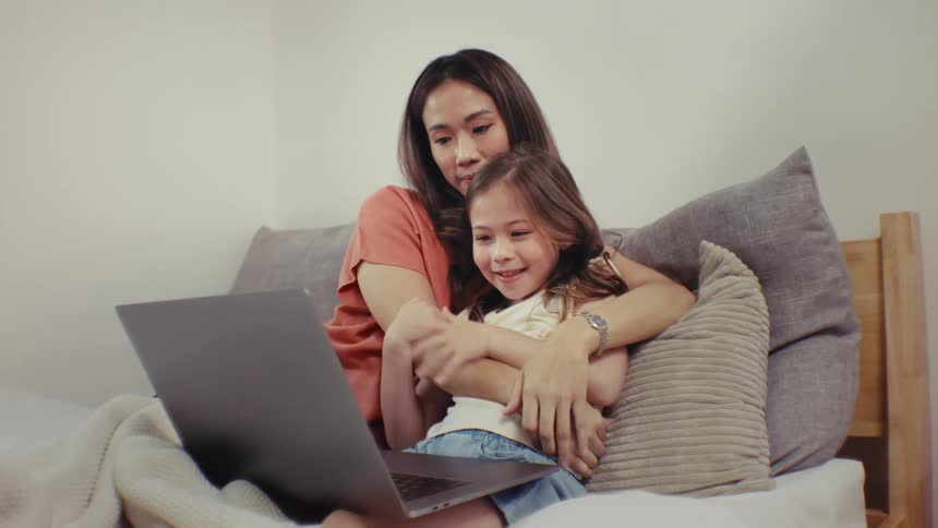 An affectionate mother and her happy young daughter cuddle together while watching a movie on a laptop in bed