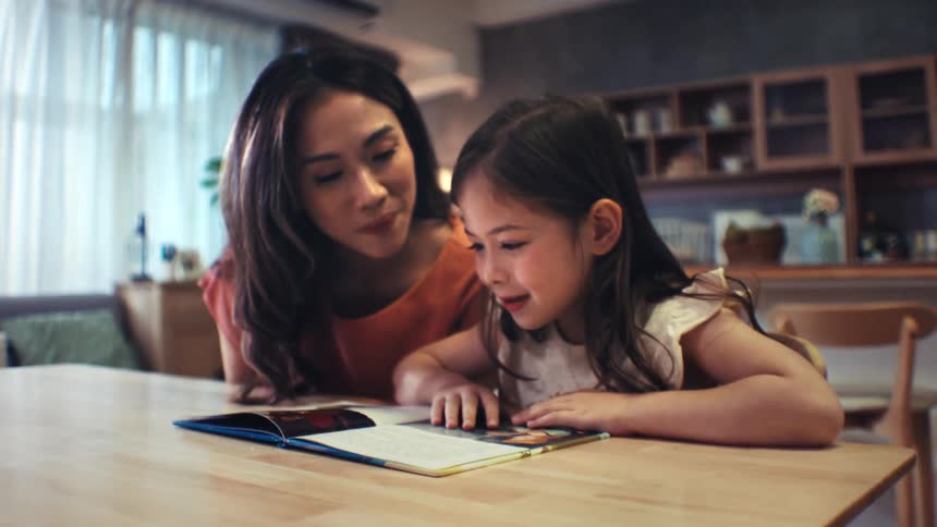 An attentive mother listens and helps as her young daughter practices reading a book aloud at the dining room table
