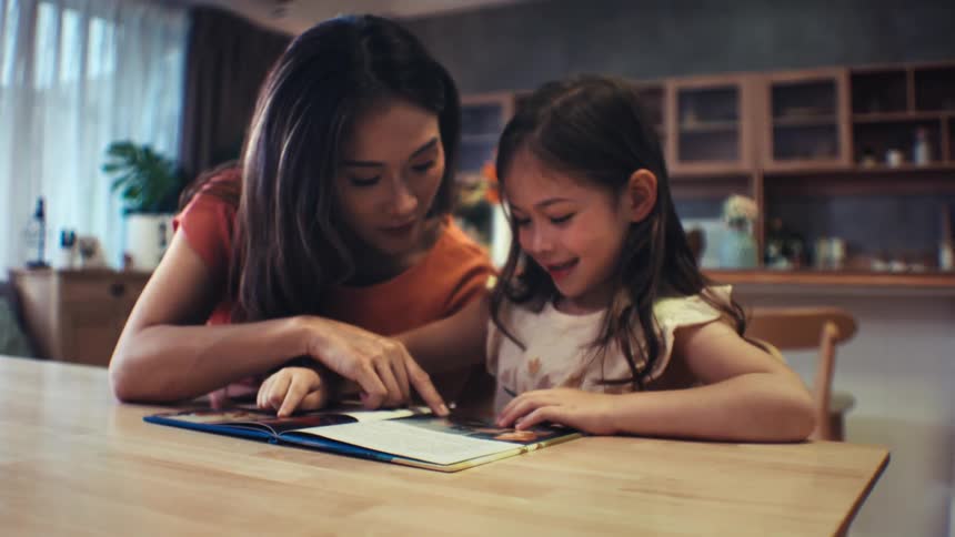 An attentive mother listens and helps as her young daughter practices reading a book aloud at the dining room table