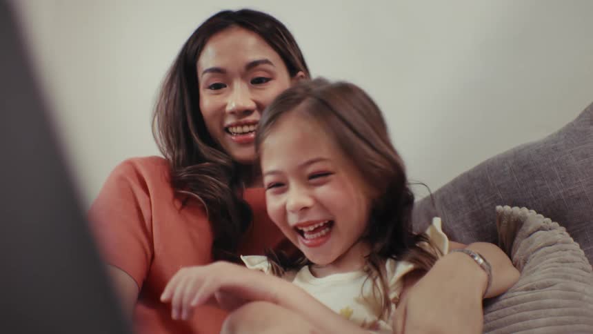 A close-up slow-motion shot of a joyful little girl laughing hysterically while watching a movie with her mother on the couch