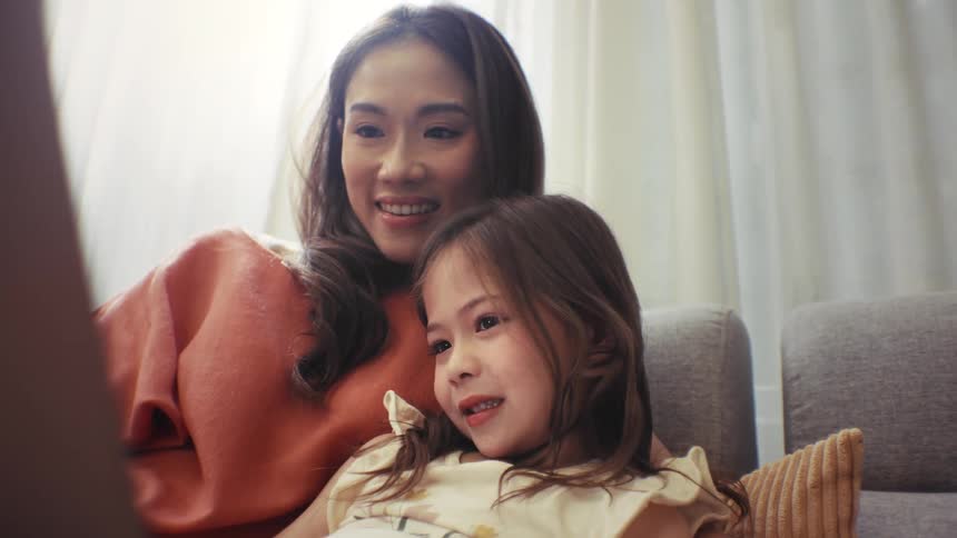Loving mother and her happy daughter share a joyful moment hugging and watching a laptop together on the sofa at home